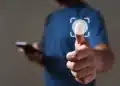 Pessoa de camiseta azul toca ícone de impressão digital em tela virtual, segurando smartphone na outra mão.