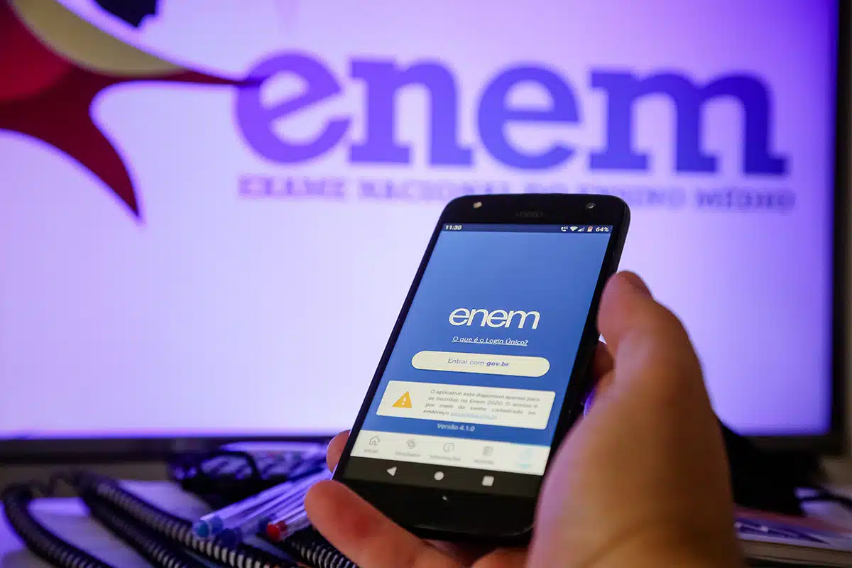 Passo a passo para entrar com recurso da isenção do Enem Celular com aplicativo do Enem aberto em frente à tela com logotipo do exame