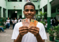 Estudante sorri segurando notas de real no pátio da escola e, ao redor, outros alunos circulam ao fundo