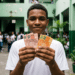Estudante sorri segurando notas de real no pátio da escola e, ao redor, outros alunos circulam ao fundo