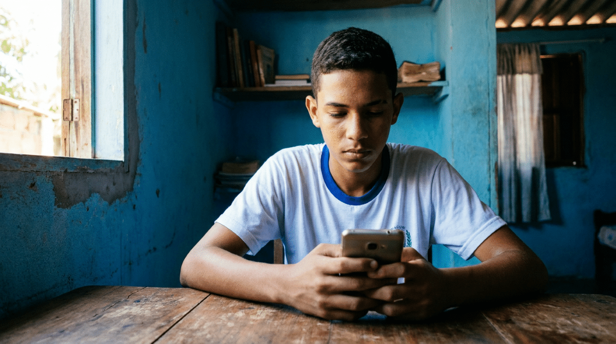 Estudante de uniforme usa o celular sentado à mesa e, ao redor, um cômodo residencial simples de paredes azuis