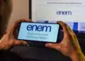Pessoa segurando celular com página do Enem aberta em frente a notebook