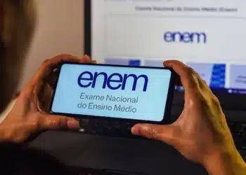 Pessoa segurando celular com página do Enem aberta em frente a notebook