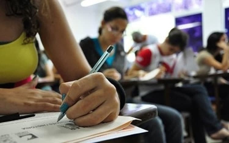Pessoas sentadas escrevendo prova em sala, jovens concentrados, cadernos e canetas em mesas