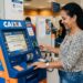 Mulher sorrindo usa caixa eletrônico da Caixa para sacar abono salarial em agência movimentada.