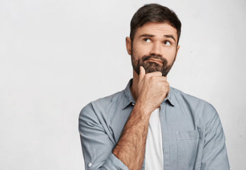 Homem jovem com barba, camisa azul claro, mão no queixo e olhar pensativo