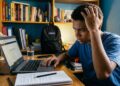 Jovem negro com mão na cabeça olha preocupado para notebook com página do Enem aberta, caderno e copo sobre mesa de estudos