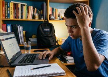 Jovem negro com mão na cabeça olha preocupado para notebook com página do Enem aberta, caderno e copo sobre mesa de estudos