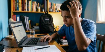 Jovem negro com mão na cabeça olha preocupado para notebook com página do Enem aberta, caderno e copo sobre mesa de estudos