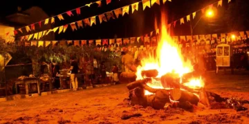 Fogueira grande acesa no centro, bandeirinhas coloridas e pessoas ao fundo em festa junina noturna