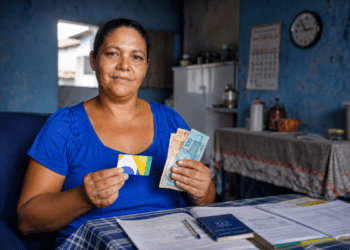 Mulher segura cartão de benefício social e cédulas de real ao lado da carteira de trabalho