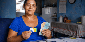 Mulher segura cartão de benefício social e cédulas de real ao lado da carteira de trabalho