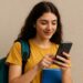 Jovem sorridente de camiseta amarela com mochila verde consulta celular segurando caderno azul