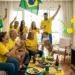 Grupo de 10 pessoas com camisas amarelas da seleção e pinturas faciais celebra gol assistindo TV com placar Brasil 2x0 Argentina