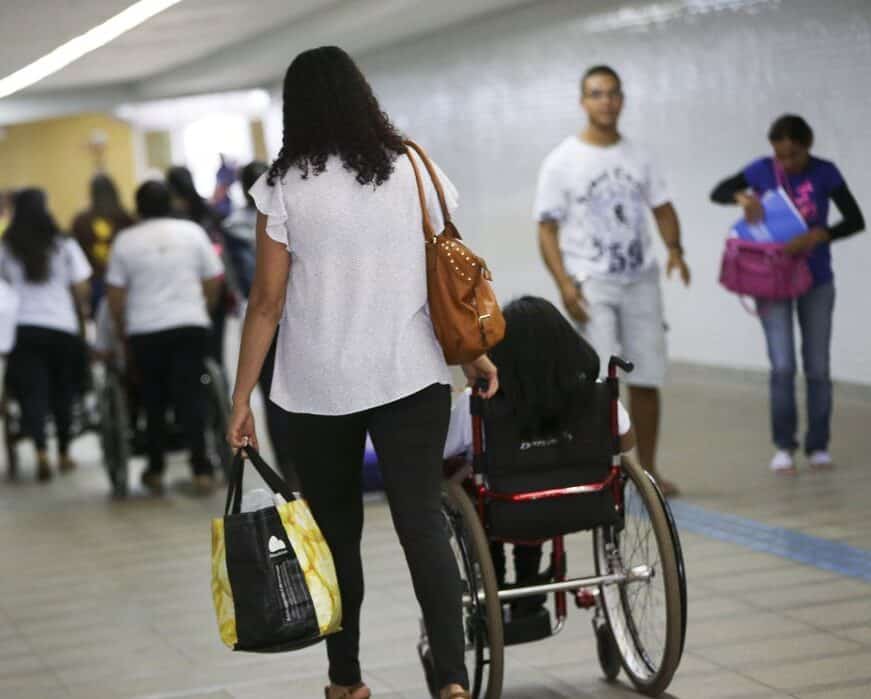 Mulher empurrando pessoa em cadeira de rodas por corredor público cercada por outros pedestres