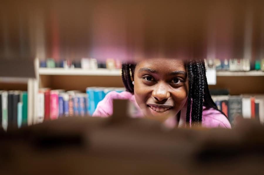 Jovem negra com tranças sorrindo entre prateleiras de livros coloridos em biblioteca, usando blusa rosa