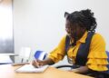 Mulher negra com tranças e óculos na cabeça, usando blusa amarela com colete azul, escreve em caderno em sala de aula
