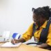 Mulher negra com tranças e óculos na cabeça, usando blusa amarela com colete azul, escreve em caderno em sala de aula