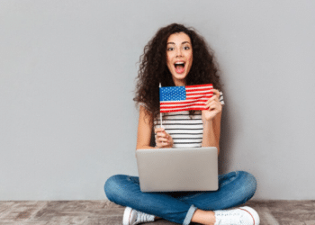 Mulher com cabelo cacheado sorri, segura bandeira americana e usa notebook sentada no chão