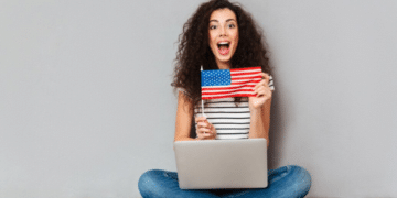 Mulher com cabelo cacheado sorri, segura bandeira americana e usa notebook sentada no chão