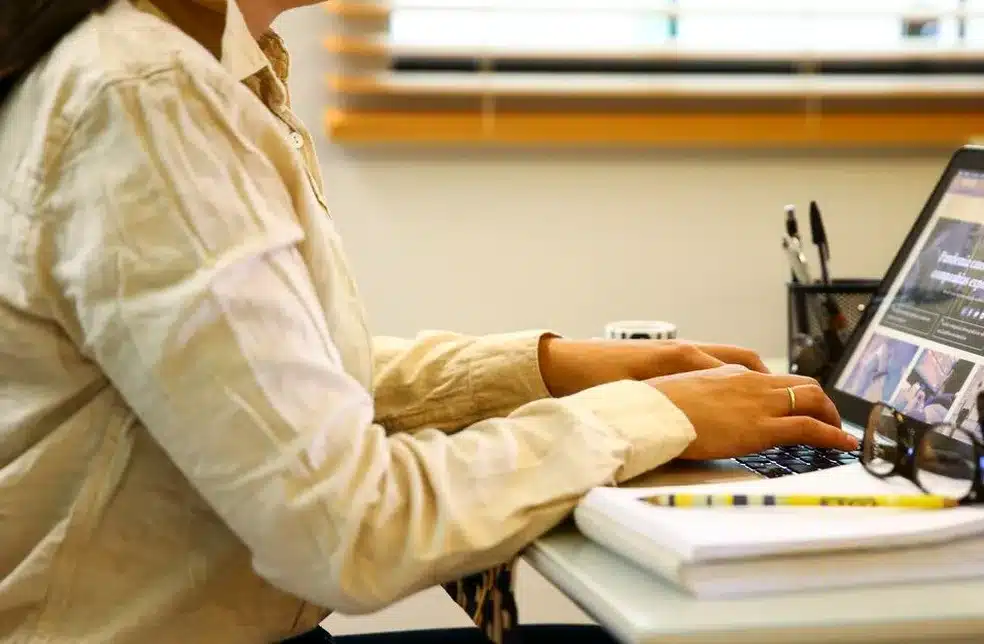 Pessoa digitando em notebook ao lado de caderno e óculos em mesa de trabalho Mulher de camisa bege digita em notebook com site aberto, ao lado de caderno, caneta e óculos em mesa de escritório