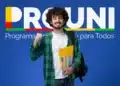 Jovem sorridente com camisa xadrez verde e mochila ergue punho à frente de banner do ProUni