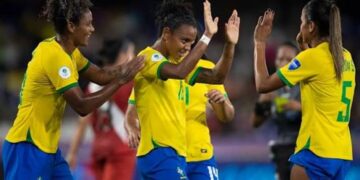 Jogadoras da seleção brasileira de futebol feminino, de uniforme amarelo e azul, comemoram juntas em campo, referência à Copa do Mundo Feminina citada no projeto