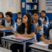 Alunos adolescentes em sala de aula, alguns atentos à explicação e outros escrevendo no caderno.