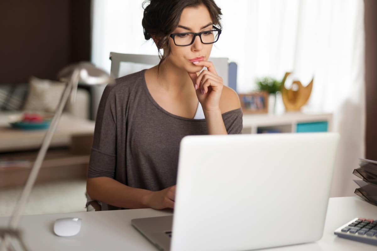 Mulher de óculos observa a tela do notebook com expressão concentrada em ambiente de trabalho em casa