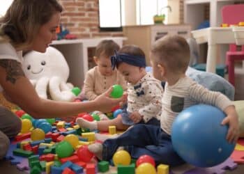 Imagem ilustrativa de educadora agachada brincando com bebês entre bolas e blocos coloridos em uma sala infantil, representando o funcionamento das Cuidotecas nas universidades
