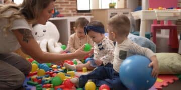 Imagem ilustrativa de educadora agachada brincando com bebês entre bolas e blocos coloridos em uma sala infantil, representando o funcionamento das Cuidotecas nas universidades