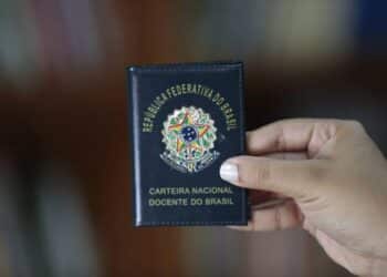 Imagem ilustrativa de mão segurando a Carteira Nacional Docente do Brasil, documento de capa azul com o brasão da República