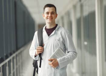 Estudante de medicina de jaleco branco e mochila segura livro em corredor de faculdade