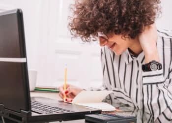 Pessoa de camisa listrada sorri enquanto escreve em caderno ao lado de um notebook aberto sobre a mesa