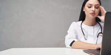Médica de jaleco branco e estetoscópio observando a tela de um notebook com expressão concentrada, cena ligada à etapa de recursos do Revalida.