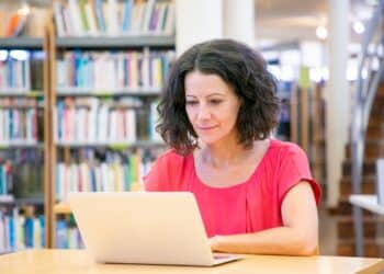 Mulher usando um notebook em uma mesa de biblioteca, com estantes de livros ao fundo