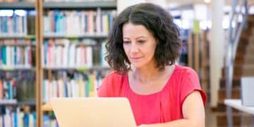 Mulher usando um notebook em uma mesa de biblioteca, com estantes de livros ao fundo