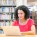 Mulher usando um notebook em uma mesa de biblioteca, com estantes de livros ao fundo