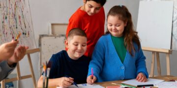 Imagem ilustrativa de três estudantes reunidos em torno de uma mesa com desenhos, lápis coloridos e tintas, em sala com cavaletes e telas ao fundo, tema da matéria sobre a nova diretriz do MEC para o ensino de arte.