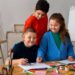 Nova diretriz do MEC consolida o ensino de arte como componente da educação básica 10 Imagem ilustrativa de três estudantes reunidos em torno de uma mesa com desenhos, lápis coloridos e tintas, em sala com cavaletes e telas ao fundo, tema da matéria sobre a nova diretriz do MEC para o ensino de arte.
