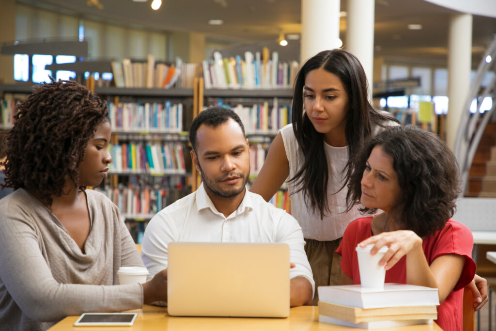 Como participar da consulta pública do MEC? Imagem ilustrativa de quatro pessoas reunidas diante de um notebook em uma biblioteca, em referência ao envio de contribuições na consulta pública do MEC