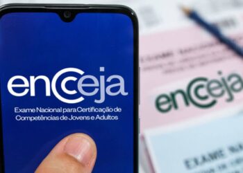 Celular exibindo a marca do Encceja, Exame Nacional para Certificação de Competências de Jovens e Adultos, com folhas de resposta e lápis desfocados ao fundo.