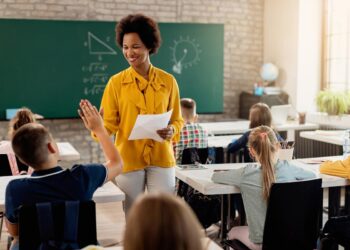 Imagem ilustrativa de professora conduzindo aula em sala com estudantes sentados e um deles com a mão levantada, em referência à consulta do MEC sobre formação continuada de professores
