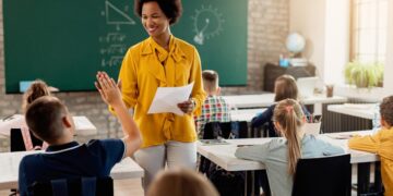 Imagem ilustrativa de professora conduzindo aula em sala com estudantes sentados e um deles com a mão levantada, em referência à consulta do MEC sobre formação continuada de professores