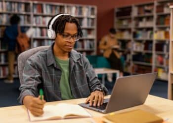 Imagem ilustrativa de jovem de fone de ouvido estudando pelo notebook e fazendo anotações em uma biblioteca, referência à modalidade de ensino a distância em debate na consulta pública
