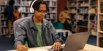 Imagem ilustrativa de jovem de fone de ouvido estudando pelo notebook e fazendo anotações em uma biblioteca, referência à modalidade de ensino a distância em debate na consulta pública