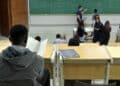 Estudante de costas lendo anotações em sala de aula universitária enquanto o professor escreve fórmulas no quadro, em imagem ligada ao programa do MEC que reestrutura a oferta de cursos.