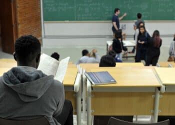 Estudante de costas lendo anotações em sala de aula universitária enquanto o professor escreve fórmulas no quadro, em imagem ligada ao programa do MEC que reestrutura a oferta de cursos.