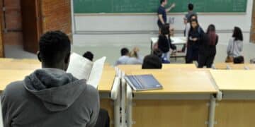 Estudante de costas lendo anotações em sala de aula universitária enquanto o professor escreve fórmulas no quadro, em imagem ligada ao programa do MEC que reestrutura a oferta de cursos.