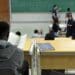 Estudante de costas lendo anotações em sala de aula universitária enquanto o professor escreve fórmulas no quadro, em imagem ligada ao programa do MEC que reestrutura a oferta de cursos.